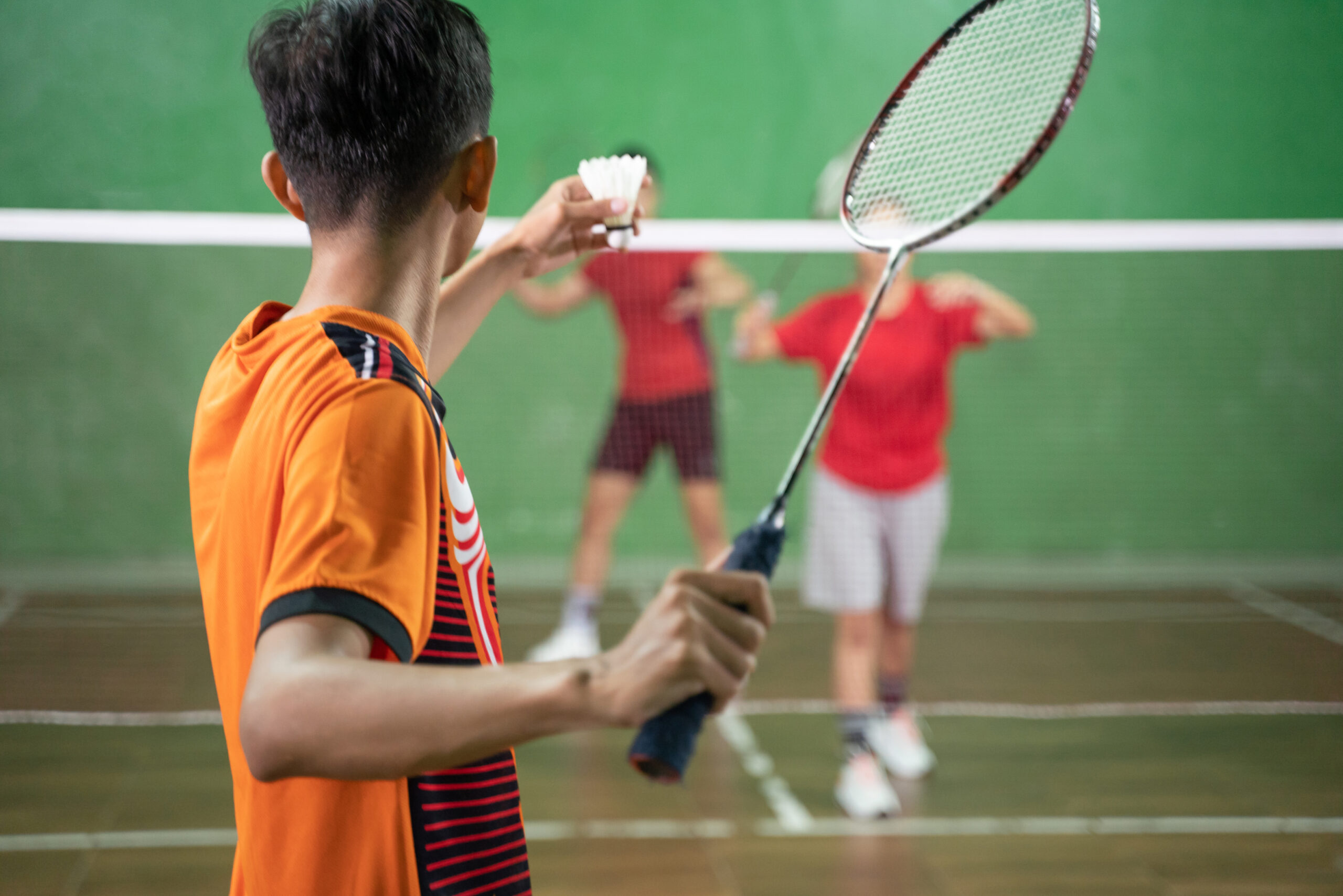 A badminton player in orange uniform holding a shuttlecock ready to serve with the opponent in a position ready to receive